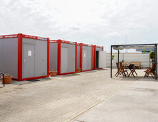 Mehrere rot-graue Container im Freien, gegenüber ein überdachter Sitzbereich.