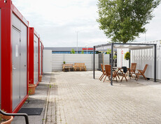Mehrere rot-graue Container im Freien, gegenüber ein überdachter Sitzbereich.