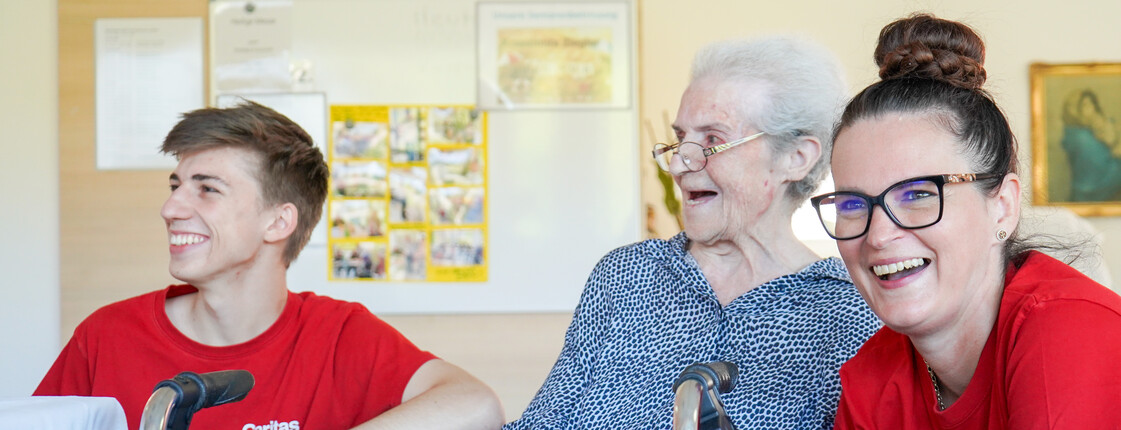 Zwei Pflegekräfte in roten Caritas-Pflege-T-Shirts, die gemeinsam mit einer Bewohnerin lachen und Zeit verbringen.